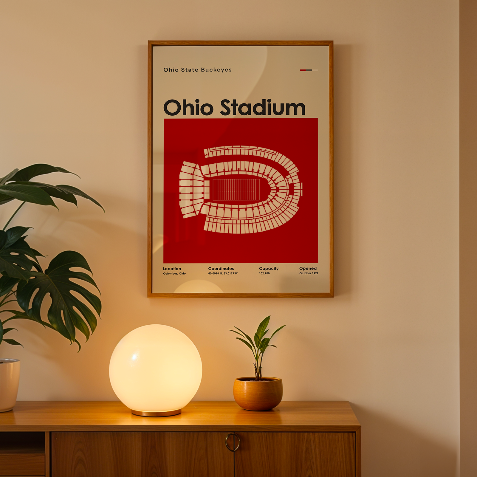 A framed Ohio Stadium poster hangs on a beige wall above a wooden console table. The poster features a bold red background with a white stadium map and the text 'Ohio Stadium.