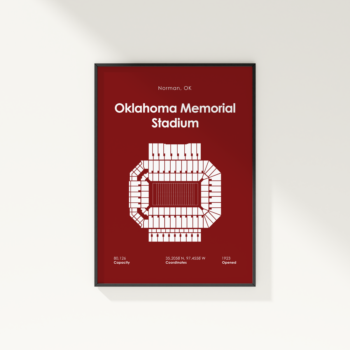 A framed vertical art print of Oklahoma Memorial Stadium in Norman, OK, featuring a white stadium floor plan and text on a dark red background, shown hanging on a light gray wall.