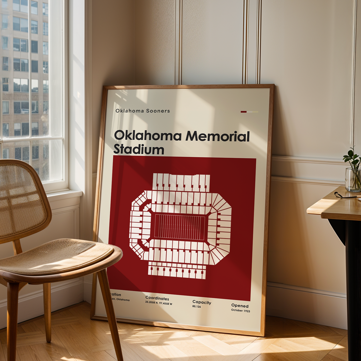 A framed Oklahoma Sooners Oklahoma Memorial Stadium poster leans against a wall in a sunlit room next to a wooden chair and a small desk.