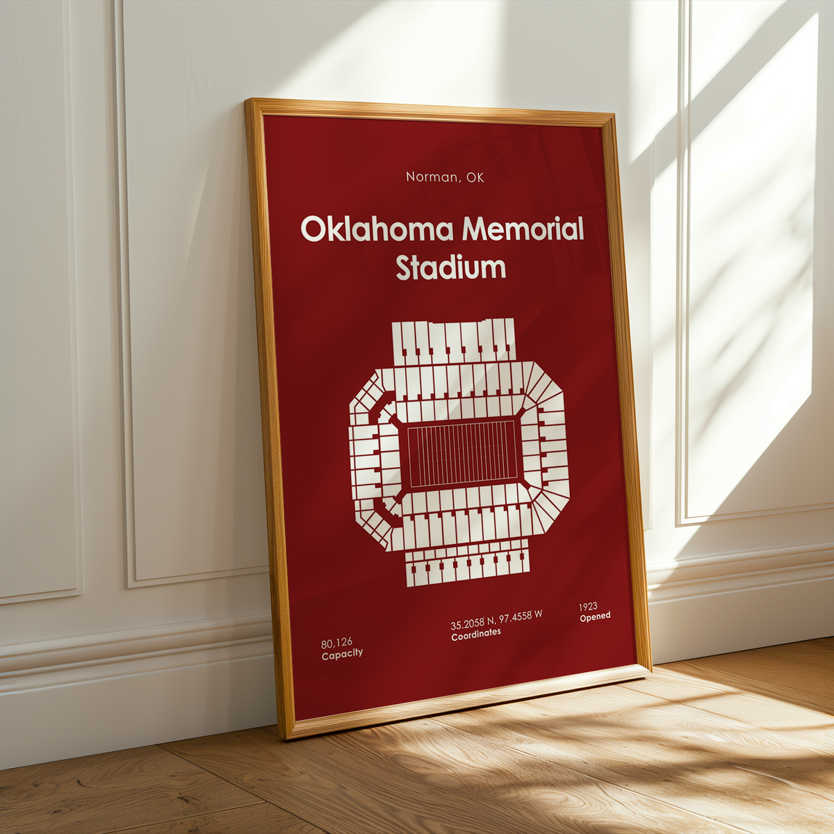 A framed Oklahoma Memorial Stadium poster leaning against a white paneled wall on a light wood floor, with warm sunlight streaming across the scene.