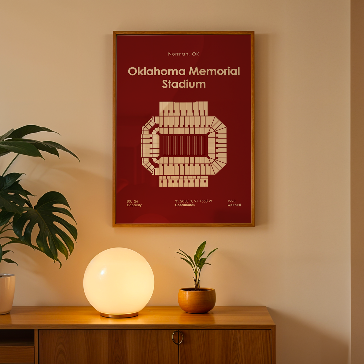 A framed Oklahoma Memorial Stadium poster hangs on a beige wall above a wooden console table. The poster is dark red with a cream stadium map and text.