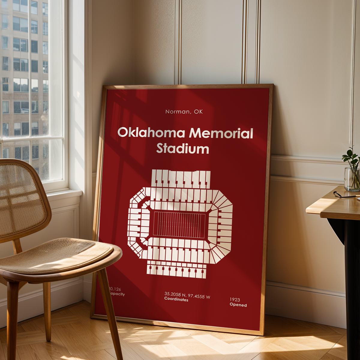 A framed poster of Oklahoma Memorial Stadium in Norman, OK, leaning against a wall in a modern room with a wooden chair and desk.