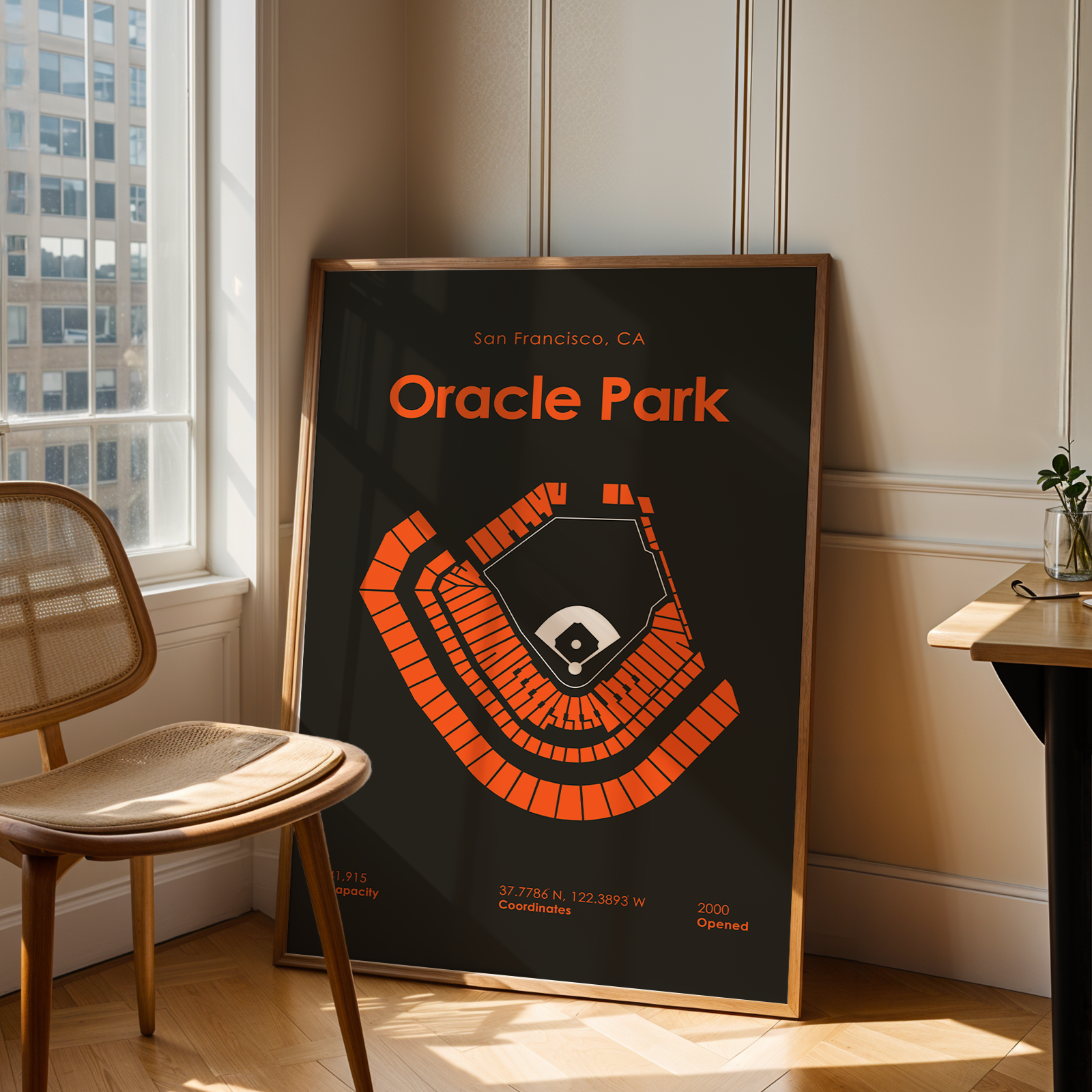 Framed poster of Oracle Park in San Francisco, CA, leaning against a wall in a room with a window and chair.