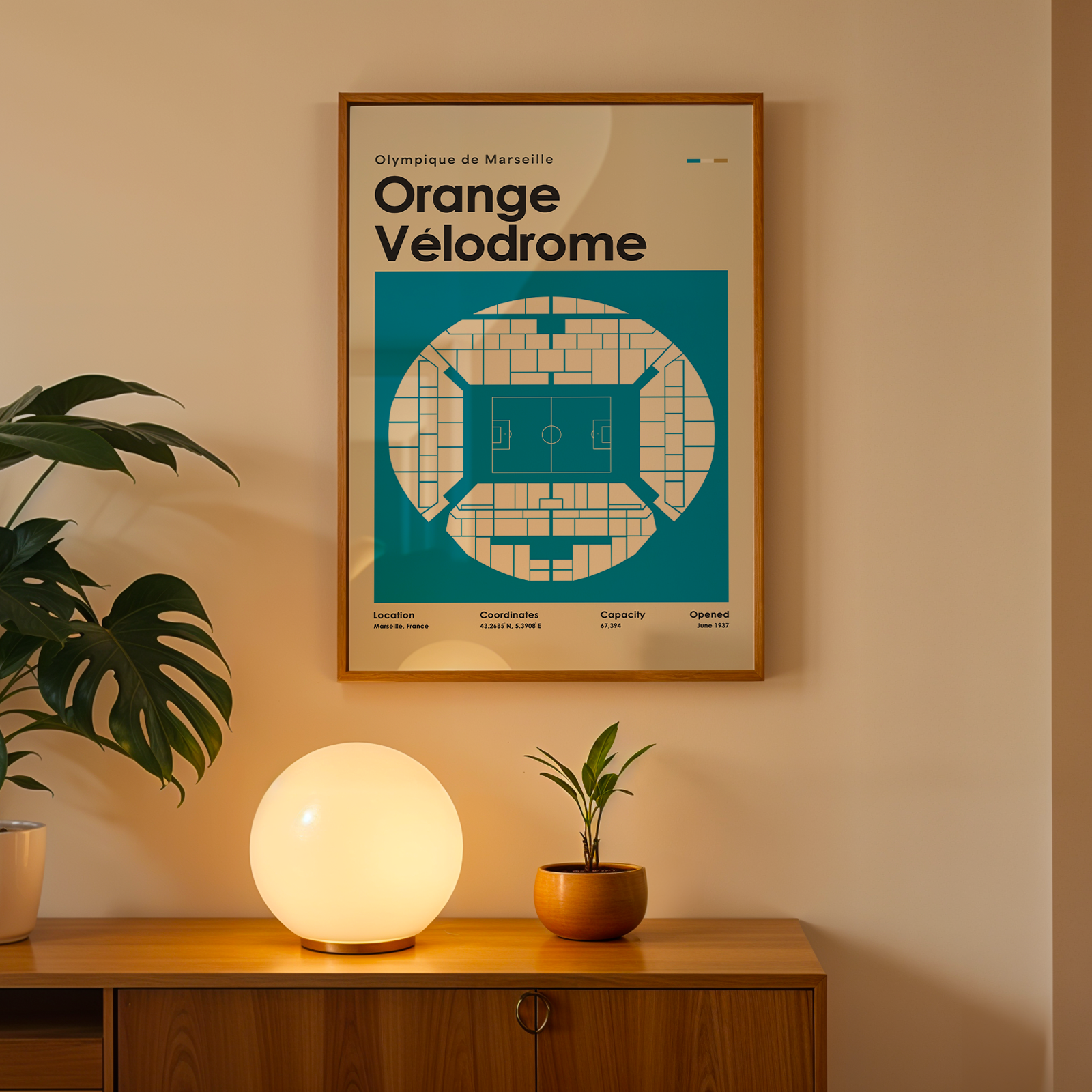 Framed poster of Orange Velodrome on a wall with a lamp and plant below.