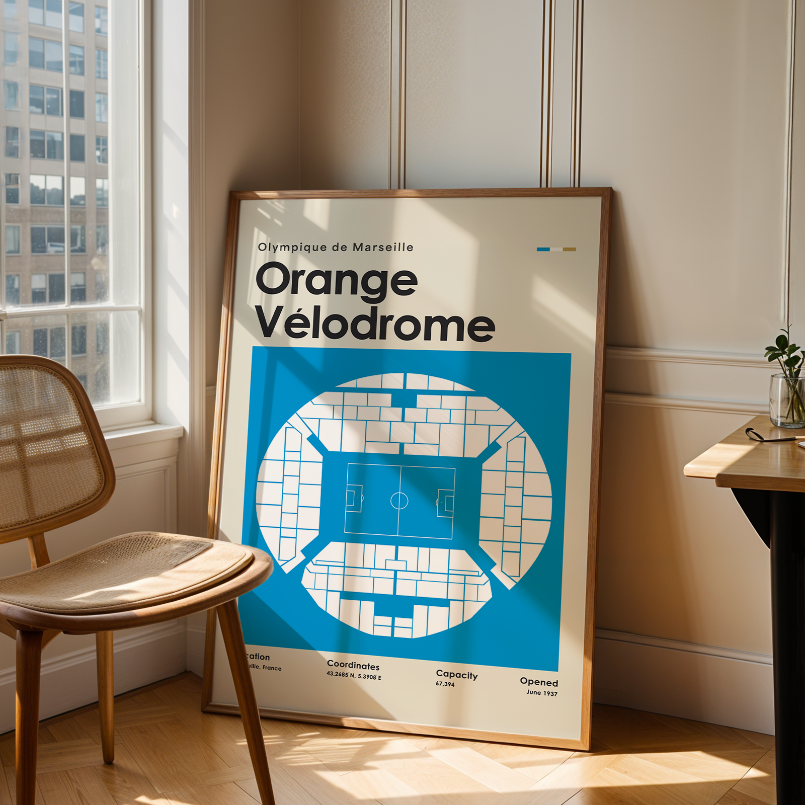 Framed poster of Orange Velodrome in a room with a chair and window.