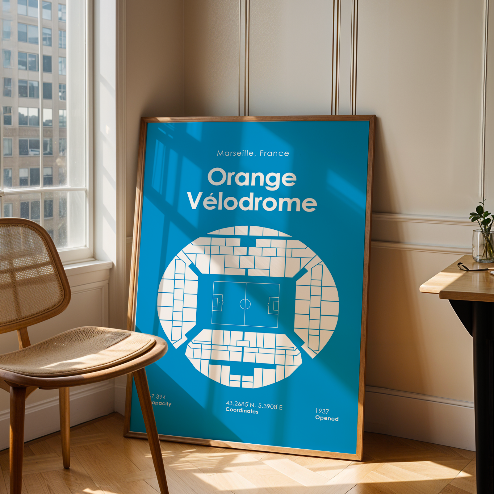 Framed poster of Orange Velodrome in Marseille, France, leaning against a wall in a room with a chair and window.