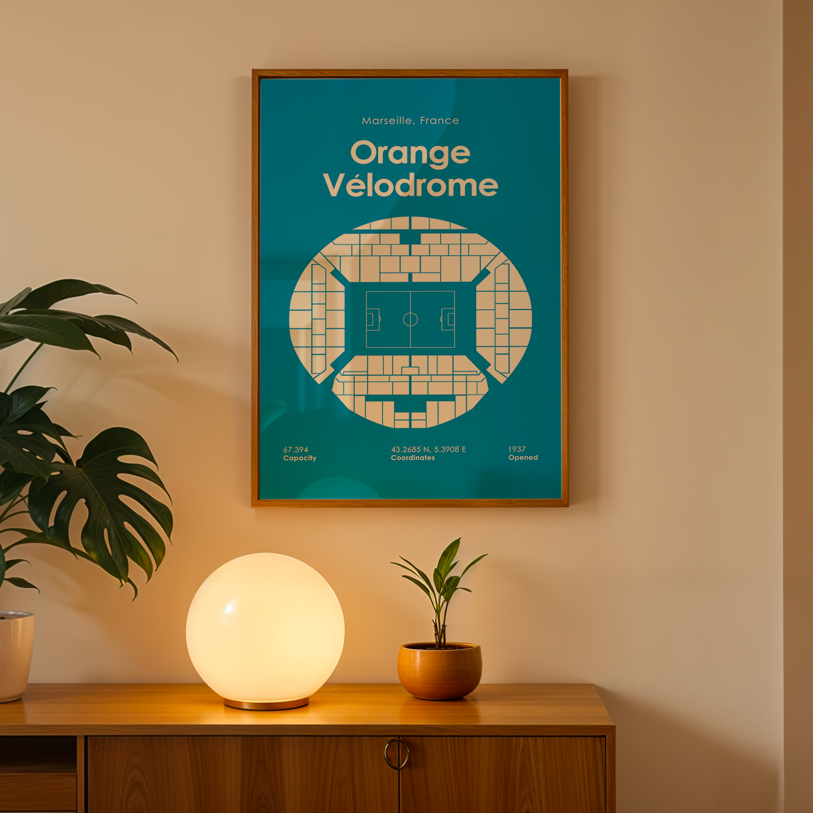 Framed poster of Orange Velodrome on a wall with a plant and lamp below.