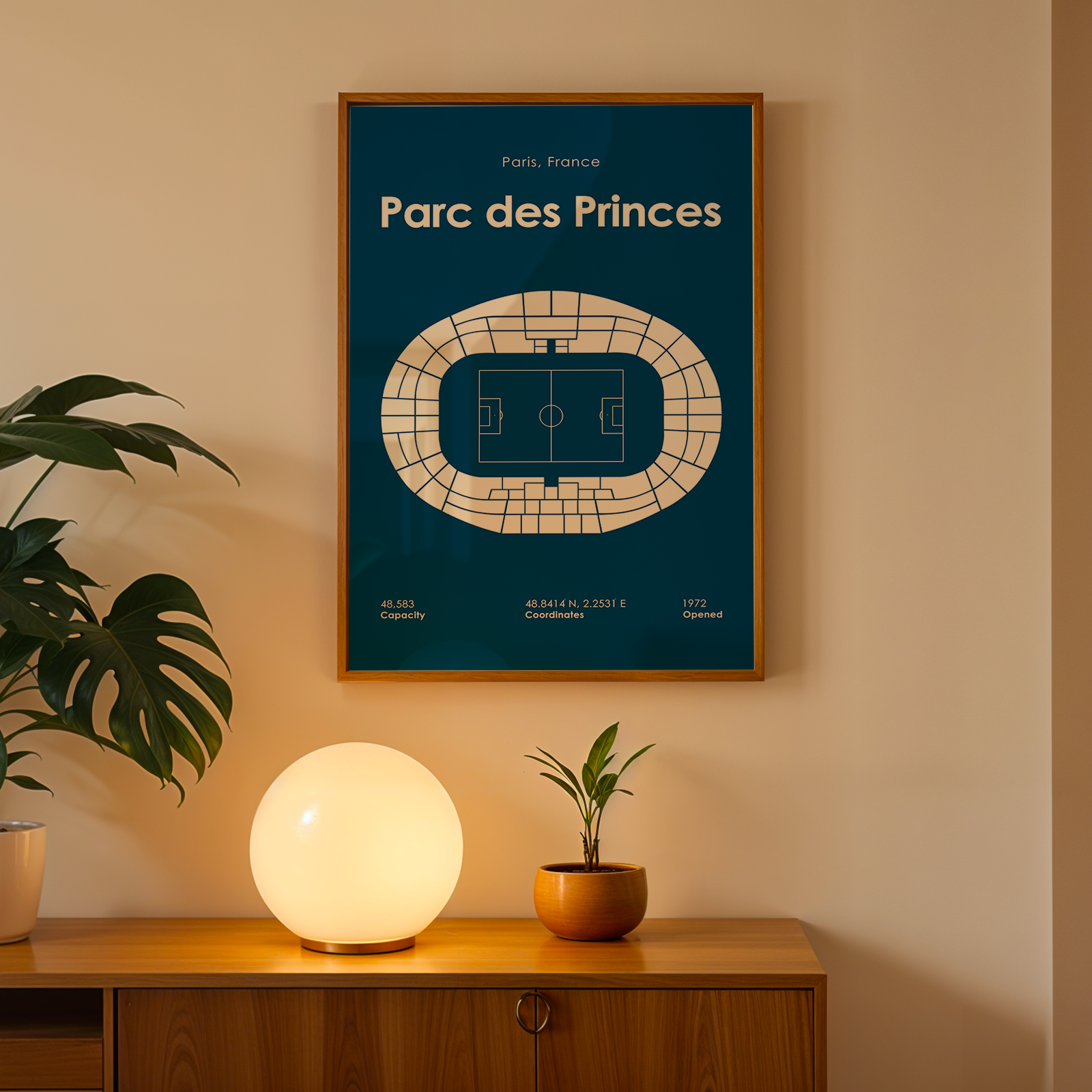 Framed poster of Parc des Princes on a wall with a lamp and plant below.