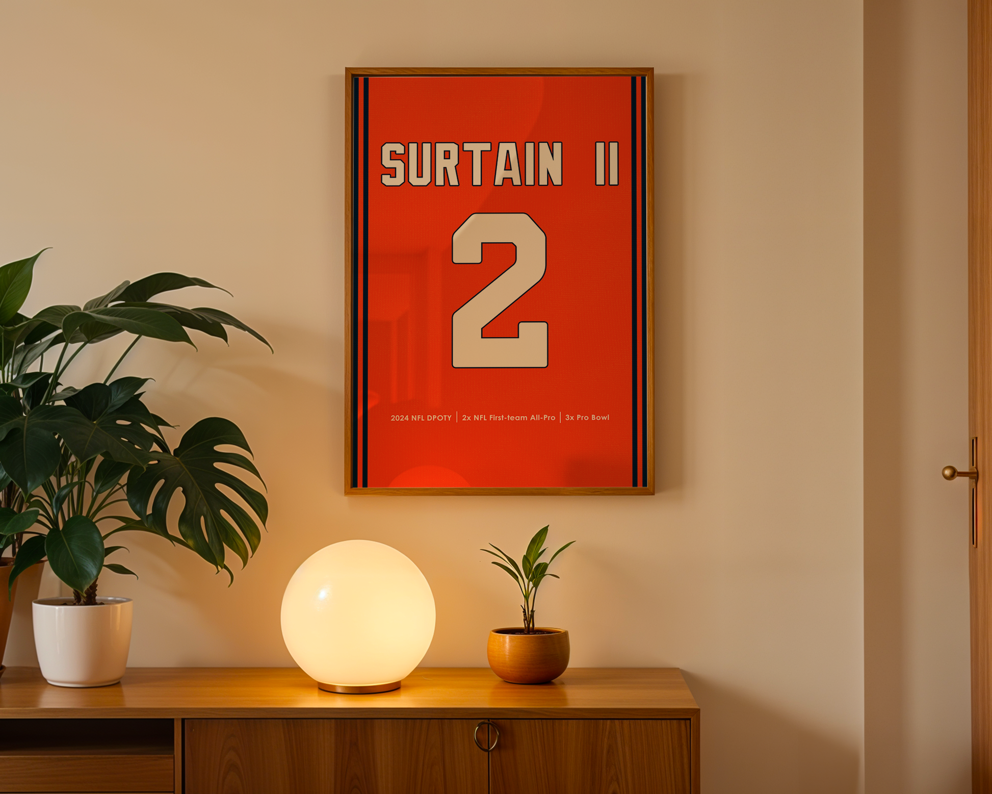 Framed artwork with 'Surtain II 2' on an orange background in a room with a plant and lamp.