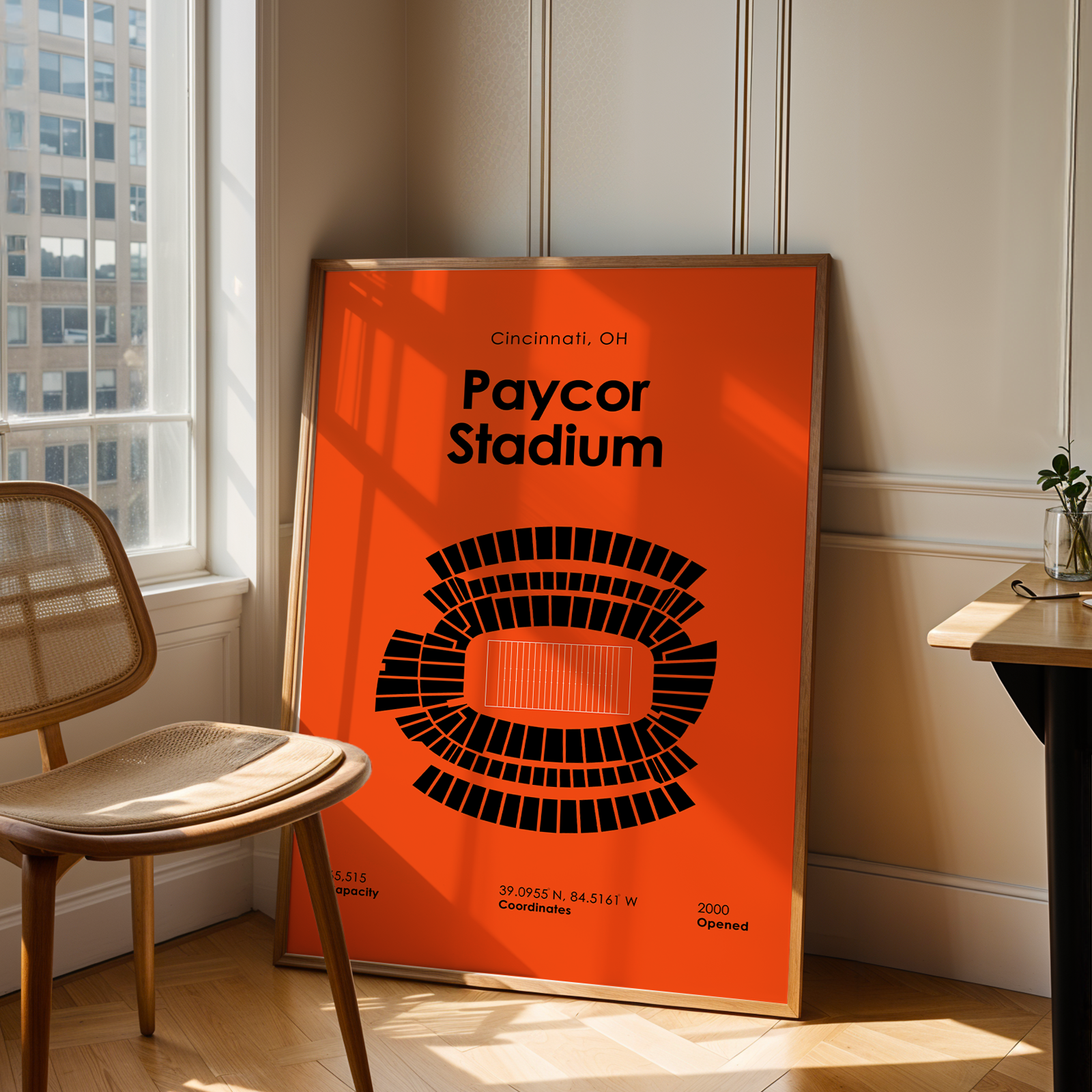 Poster of Paycor Stadium in an orange frame leaning against a wall next to a chair and table.