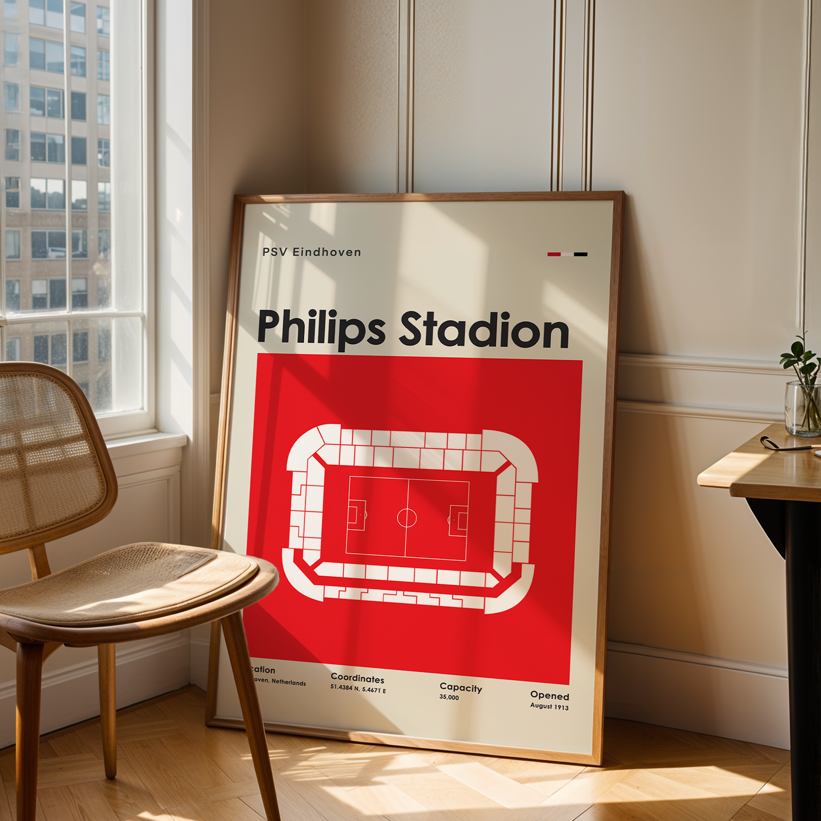 A framed Philips Stadion sports poster leaning against a wall in a sunlit room, next to a wooden chair and a small desk with a plant.