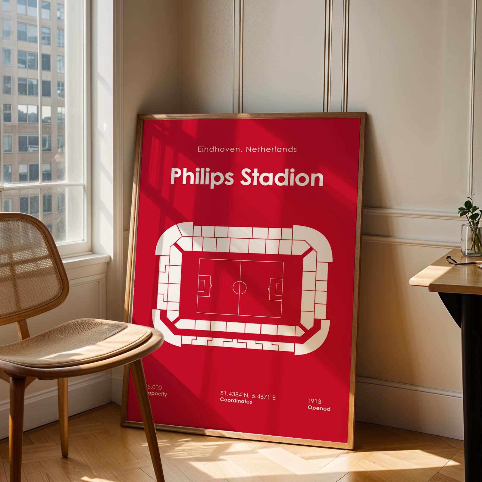 A framed red and white Philips Stadion sports poster leaning against a wall in a sunlit room next to a wooden chair and desk.