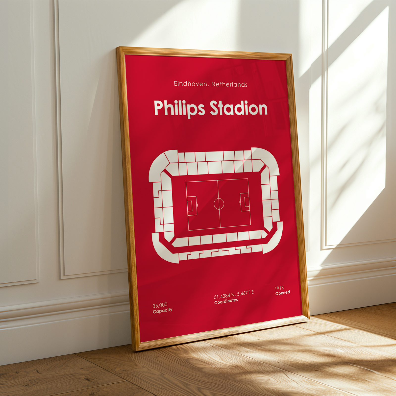 A framed sports poster of Philips Stadion in Eindhoven, Netherlands, leaning against a white paneled wall on a wooden floor with warm sunlight streaming in from a window.