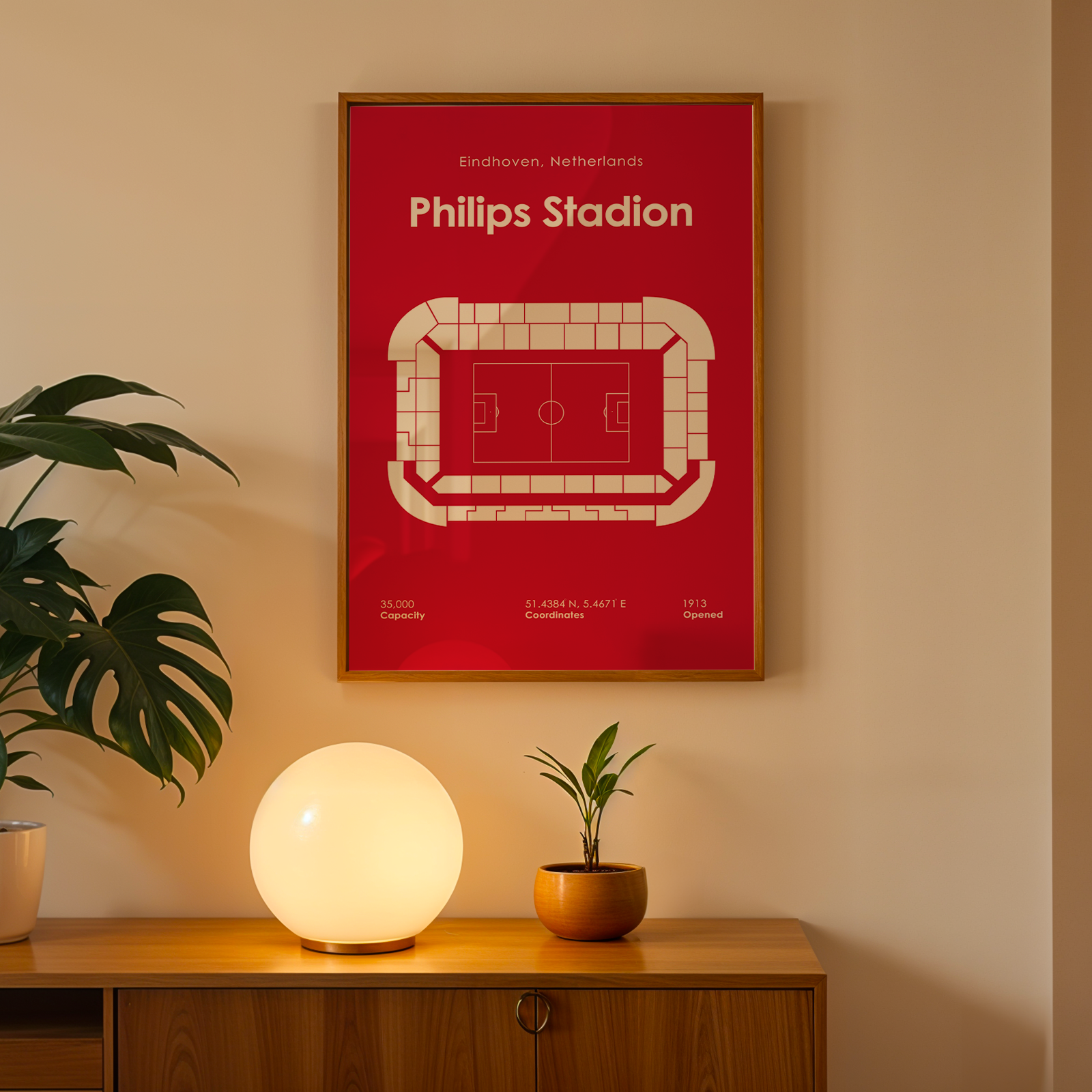 A framed Philips Stadion football stadium print in red and white hangs on a beige wall above a wooden console table with a glowing globe lamp and potted plants.