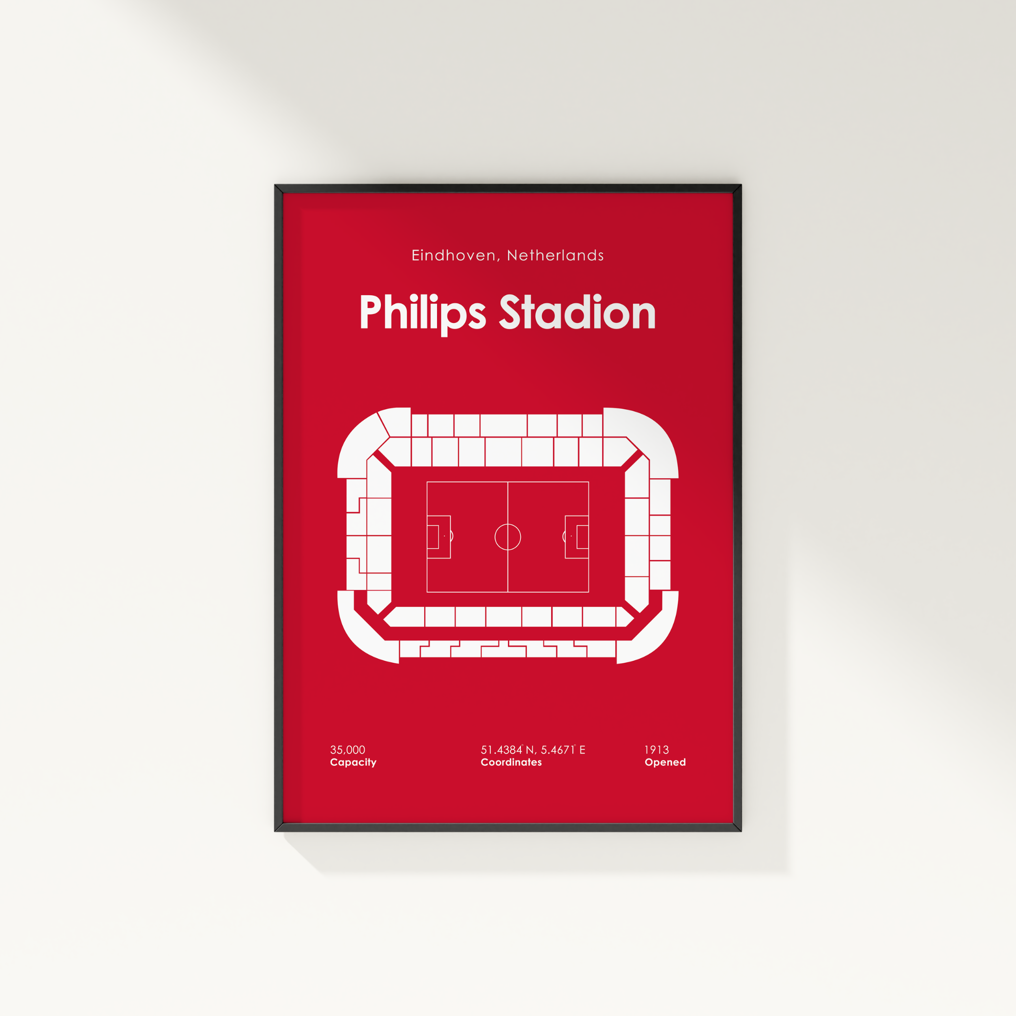 A framed minimalist stadium seating poster for Philips Stadion in Eindhoven, Netherlands, featuring a bold red background and white line art of the pitch and stands, shown hanging on a light gray wall.