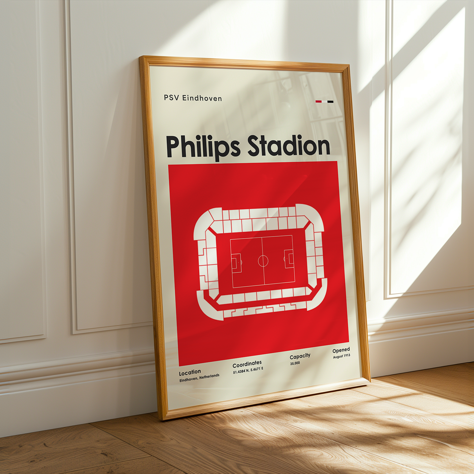 A framed PSV Eindhoven Philips Stadion sports poster leaning against a white paneled wall on a light wood floor, with warm sunlight casting shadows across the scene.