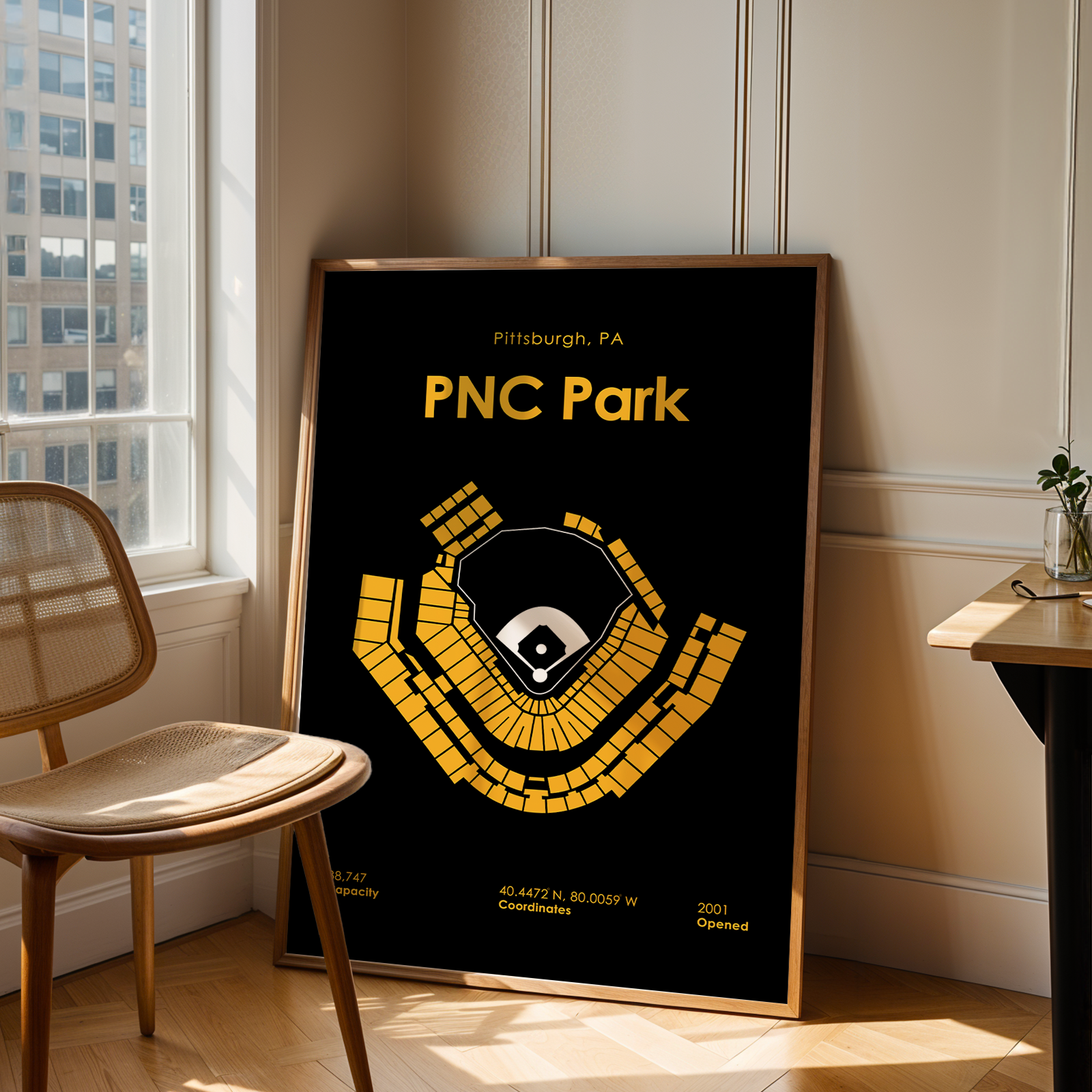 Framed poster of PNC Park in Pittsburgh, PA on a wall in a room with a window and chair.