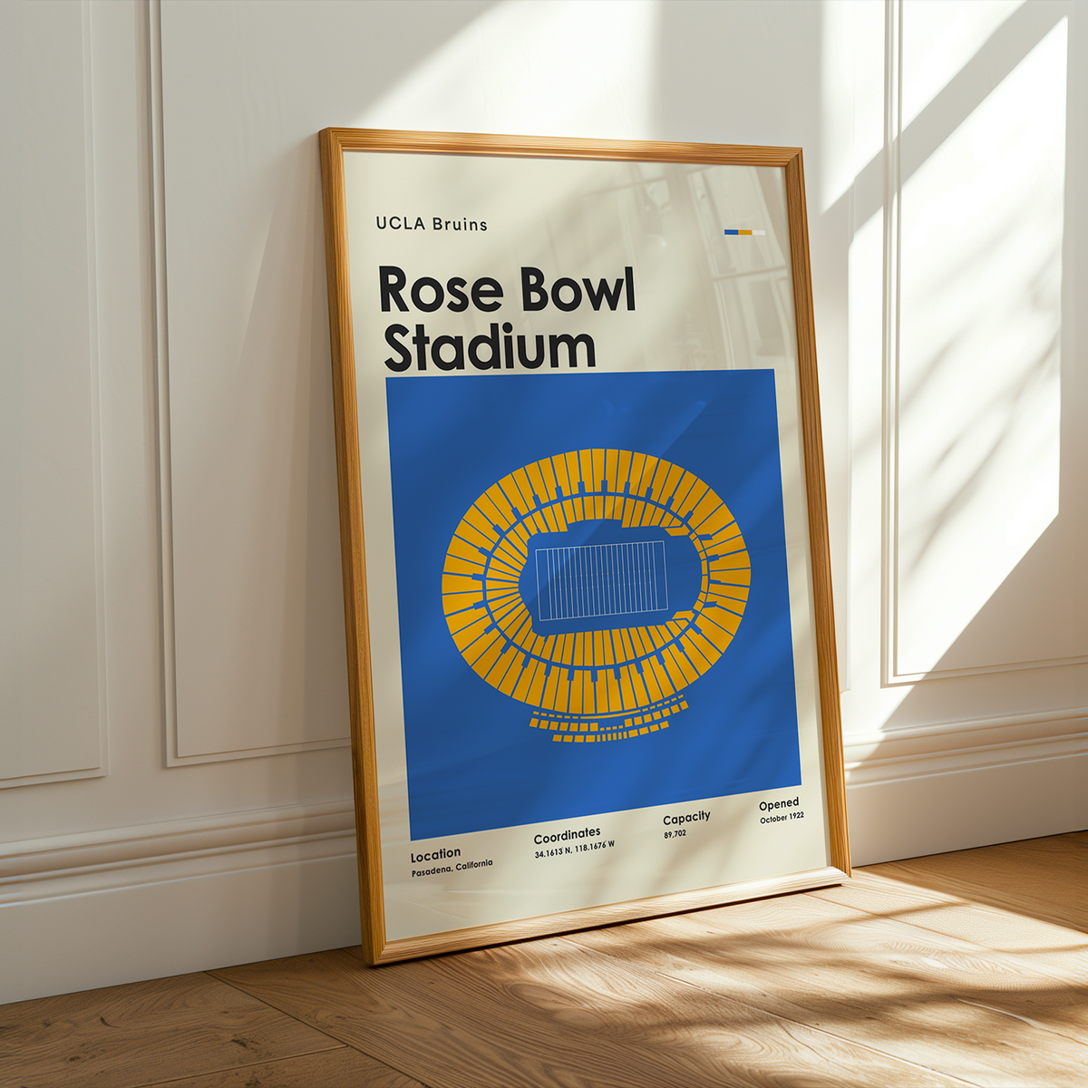 A framed poster of UCLA Bruins Rose Bowl Stadium leaning against a white paneled wall on a wooden floor, with warm sunlight casting shadows across the scene.