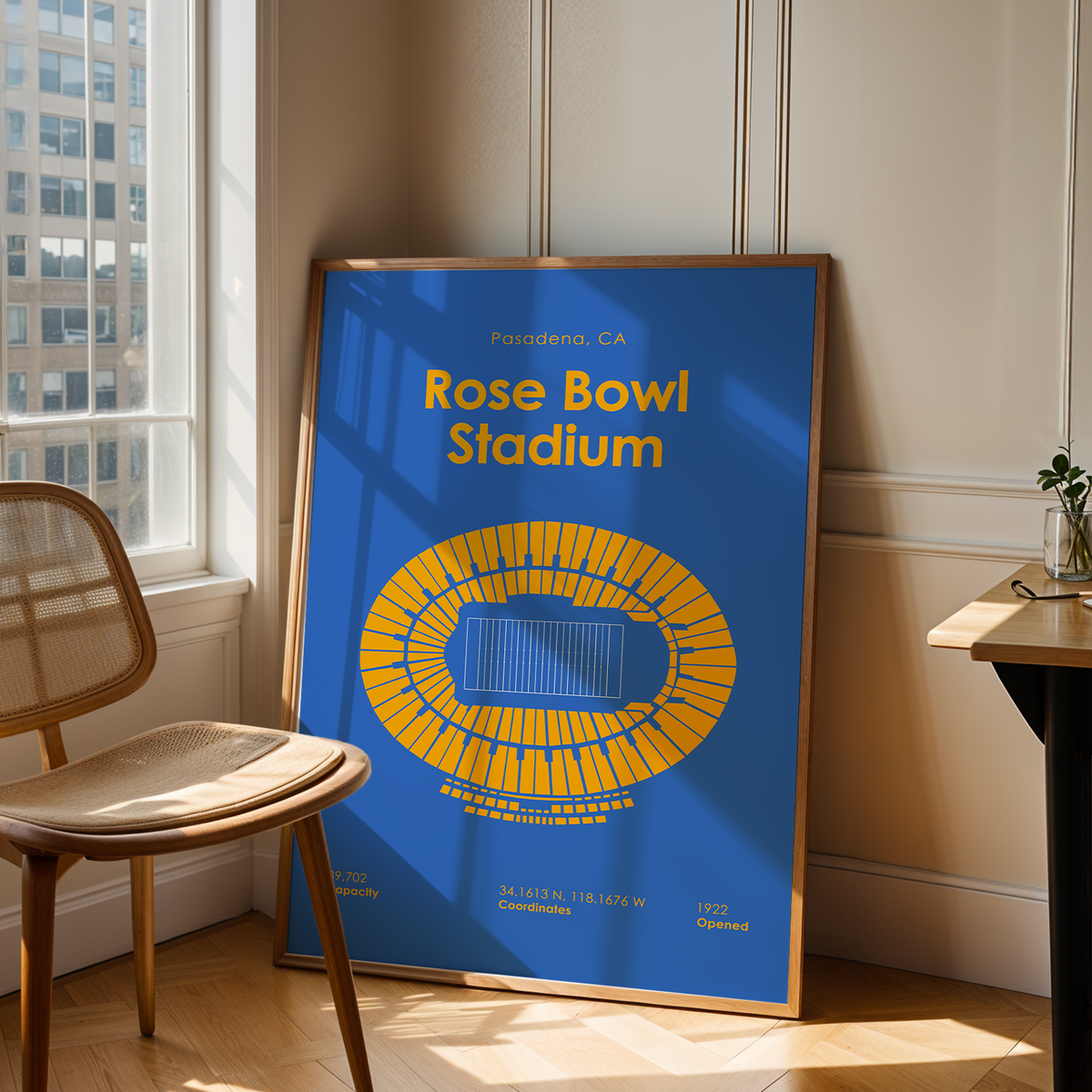A framed poster of Rose Bowl Stadium in Pasadena, CA, leaning against a wall in a sunlit room next to a wooden chair and desk.