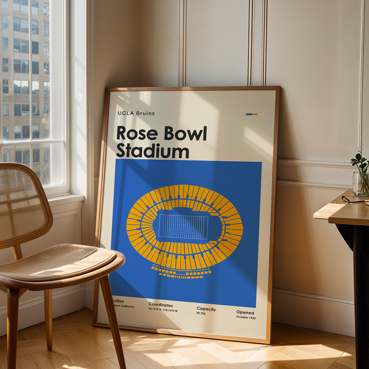 A framed poster of UCLA Brains Rose Bowl Stadium leaning against a wall in a sunlit room next to a wooden chair and desk.