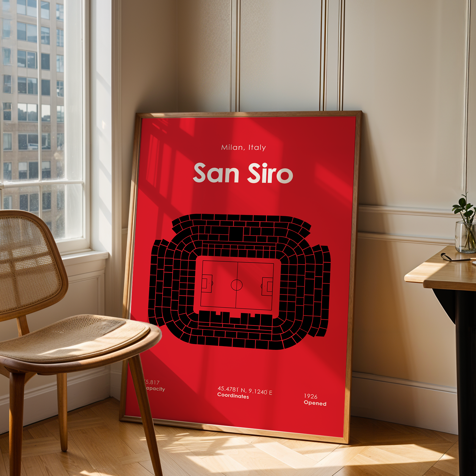 Framed poster of San Siro stadium layout on a red background in a room with a window and chair.