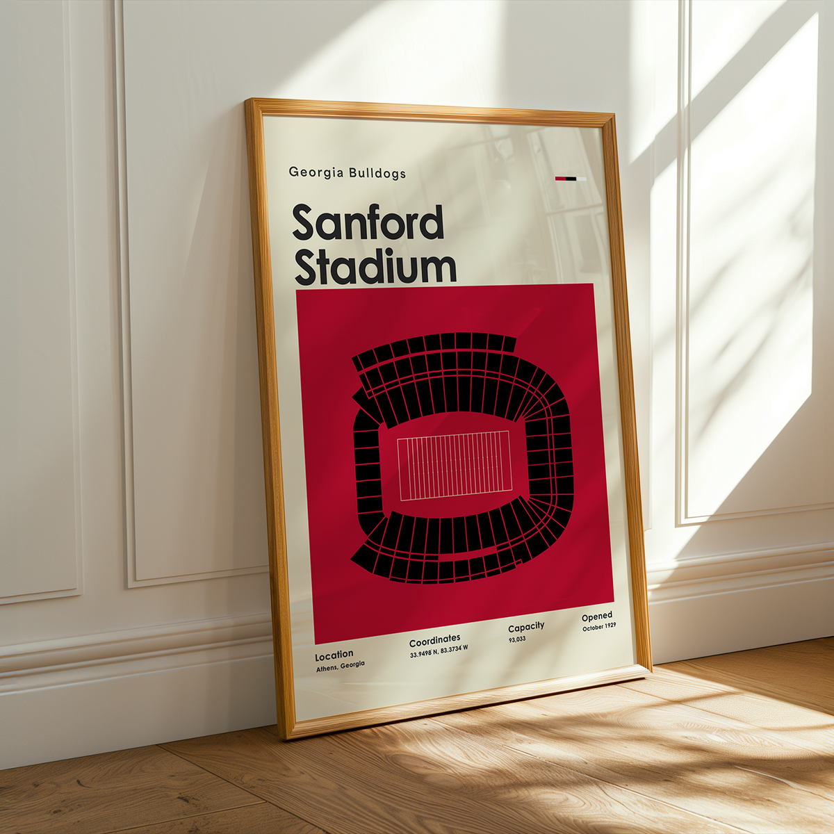 A framed Georgia Bulldogs Sanford Stadium poster leans against a white paneled wall on a light wood floor, with warm sunlight casting shadows across the scene.