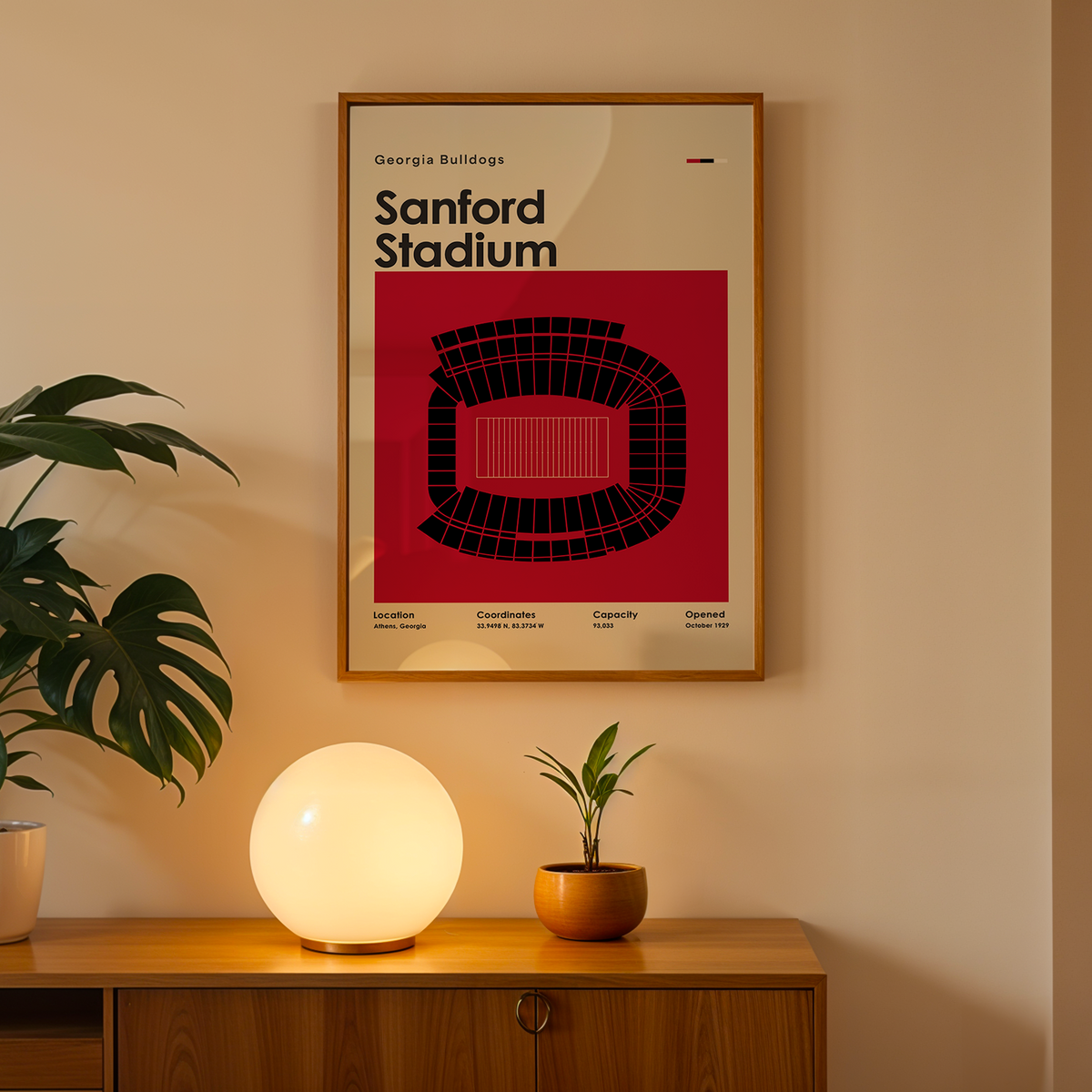 A framed poster of Sanford Stadium by Georgia Bulldogs hanging on a beige wall above a wooden console table. The poster features a bold red background with a black stadium graphic and black text.
