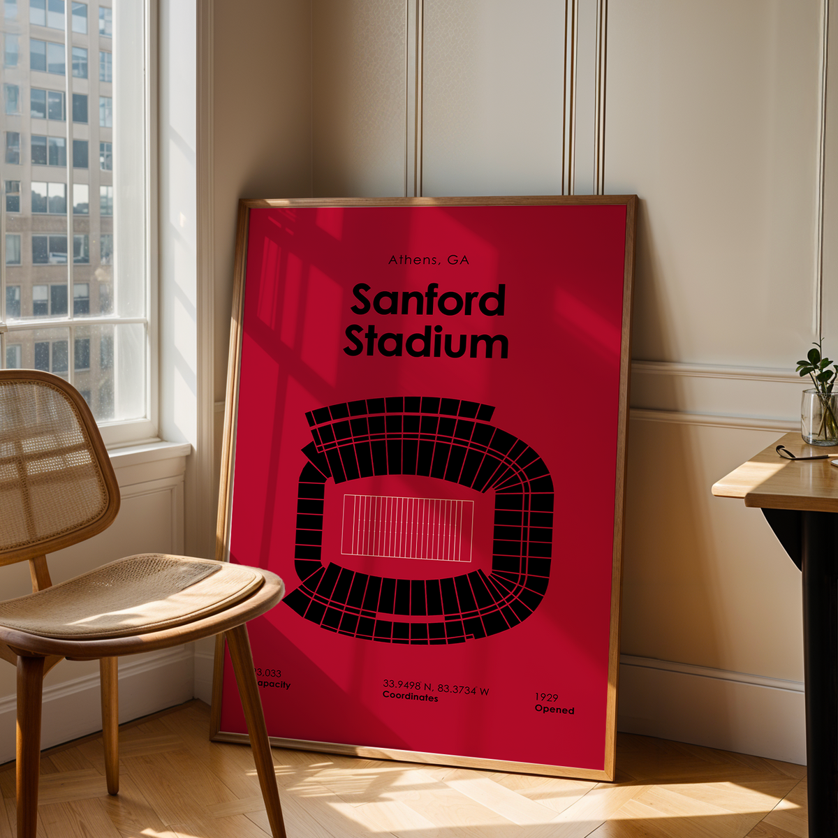 A framed poster of Sanford Stadium in Athens, GA, leaning against a wall in a modern room with a wooden chair and a desk.