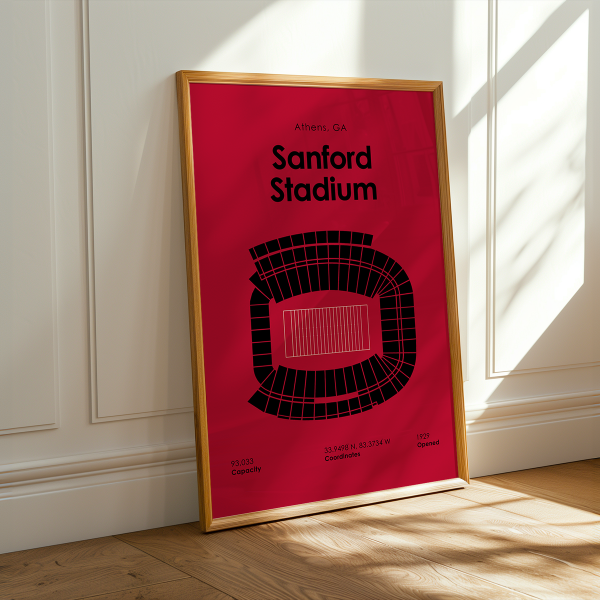 A framed minimalist poster of Stanford Stadium in Athens, GA, leaning against a white paneled wall on a light wood floor with warm sunlight and window shadows.