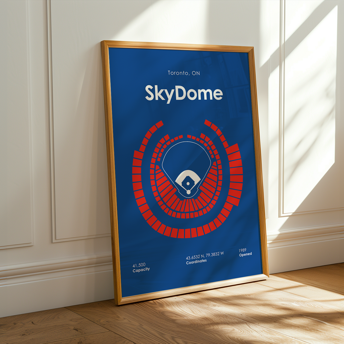 A framed poster of the Toronto SkyDome baseball stadium, leaning against a white paneled wall on a wooden floor with warm sunlight streaming in from a window.