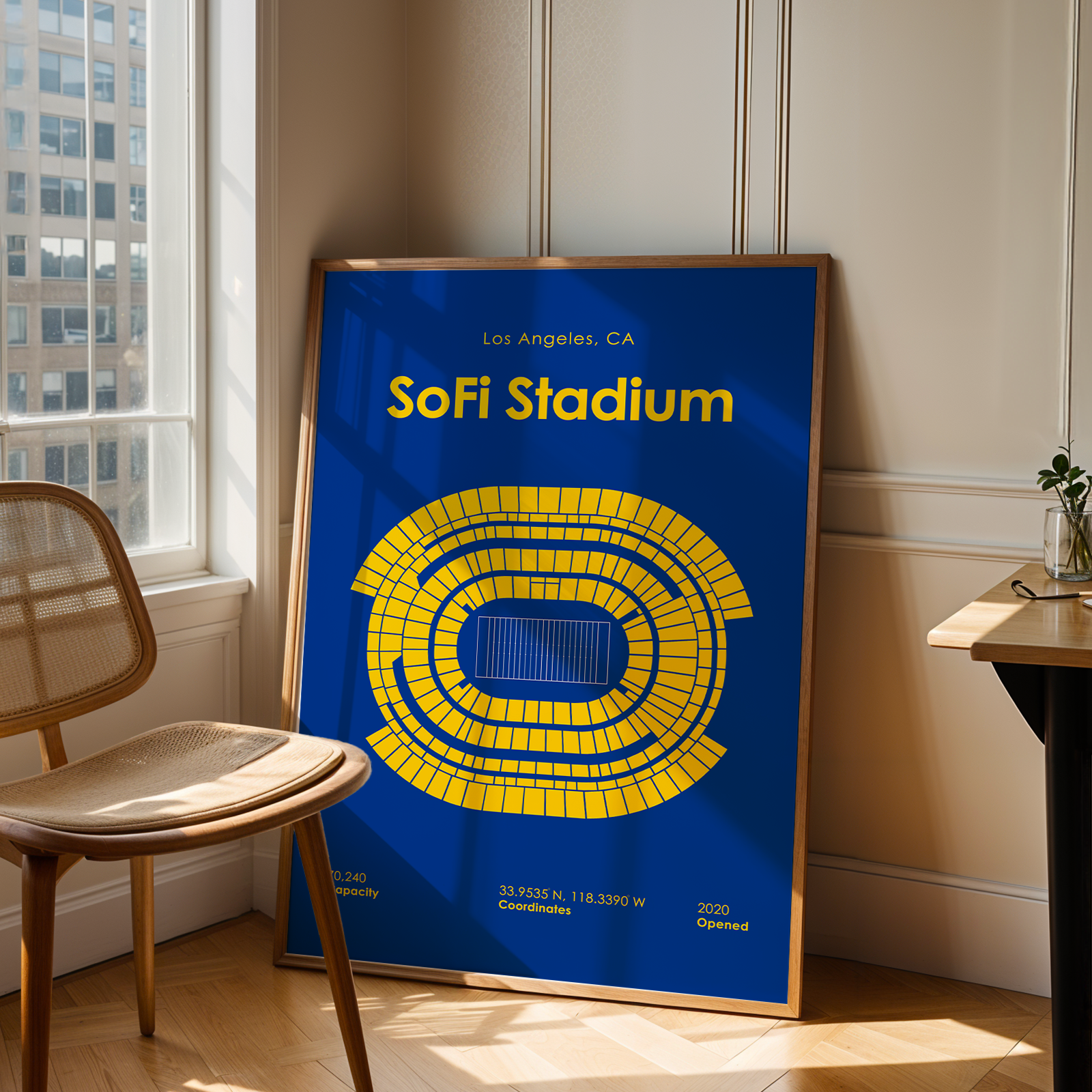 Framed poster of SoFi Stadium in Los Angeles, CA, leaning against a wall in a room with a chair and table.