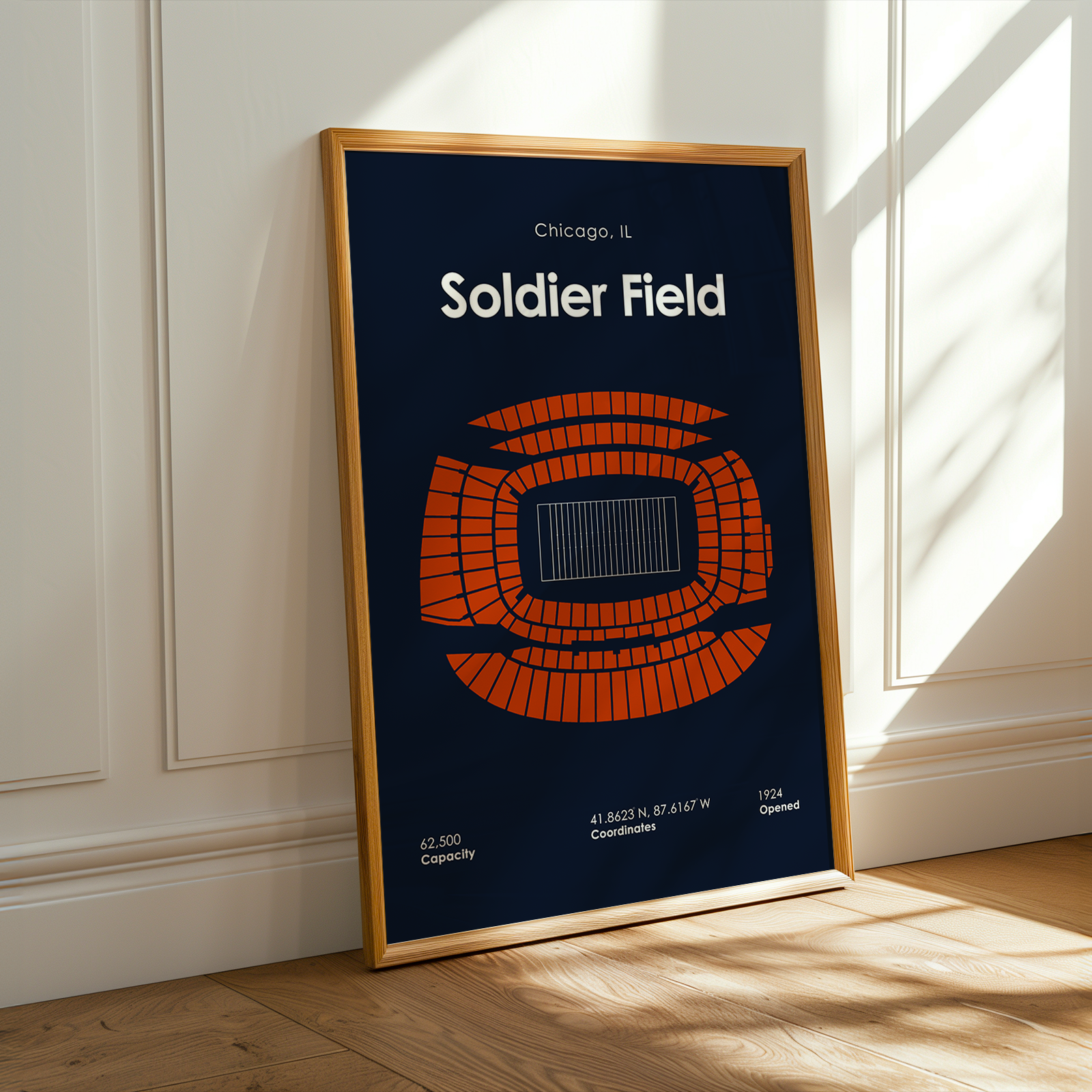 Framed poster of Soldier Field with a blue background and orange seating diagram, leaning against a white wall.