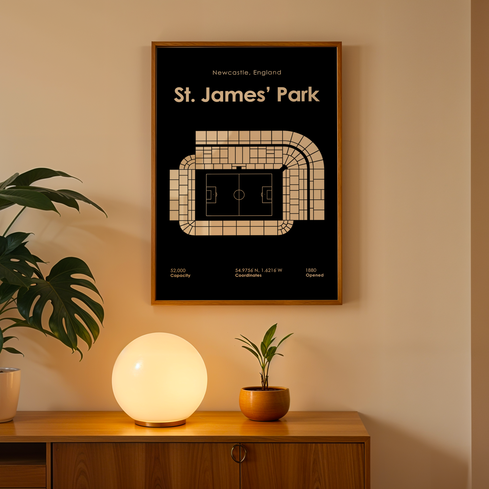 Framed poster of St. James' Park stadium on a wall with a lamp and plant below.