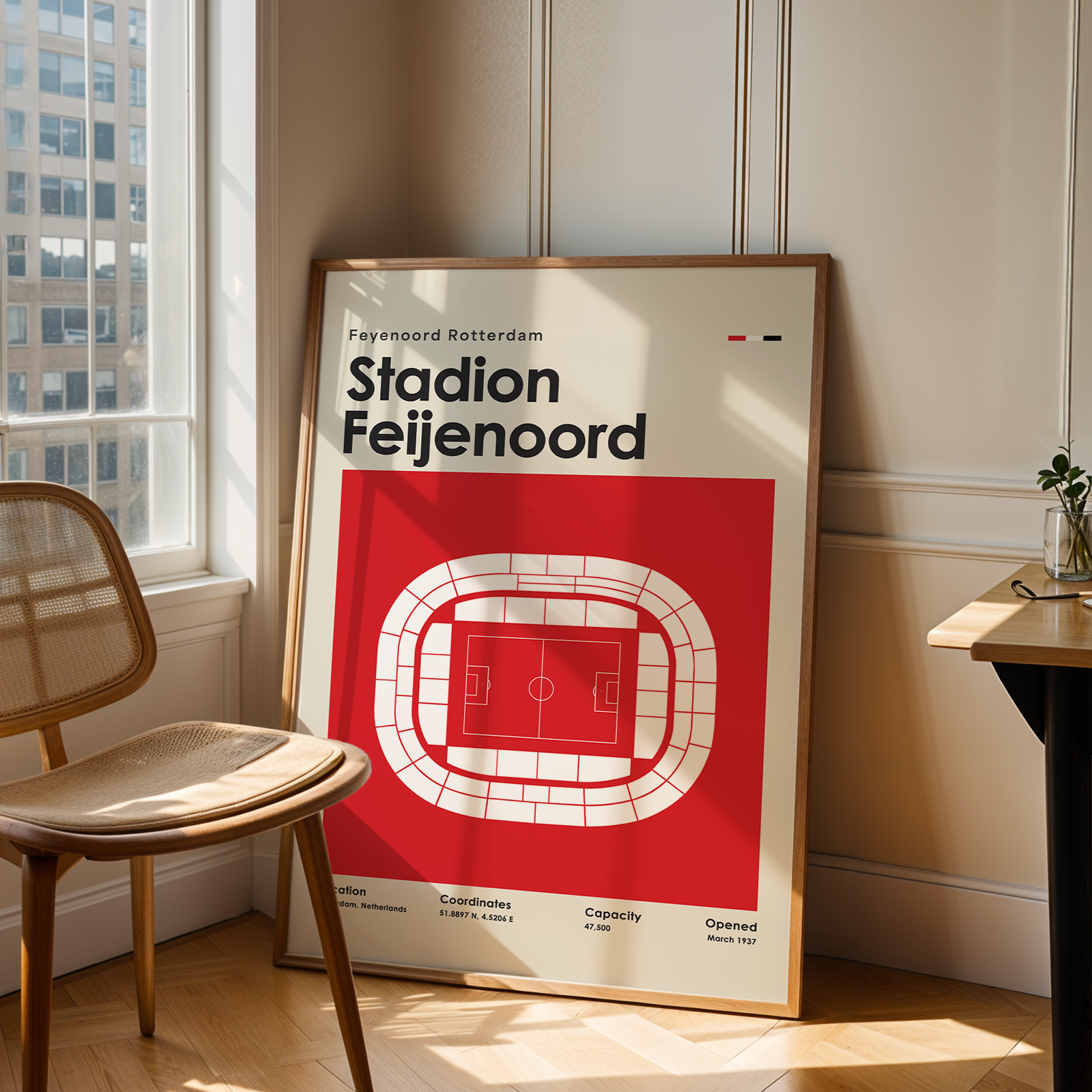 A framed vintage-style football stadium poster for Feyenoord Rotterdam / Stadion Feijenoord leans against a wall in a sunlit room beside a wooden chair and a small desk.