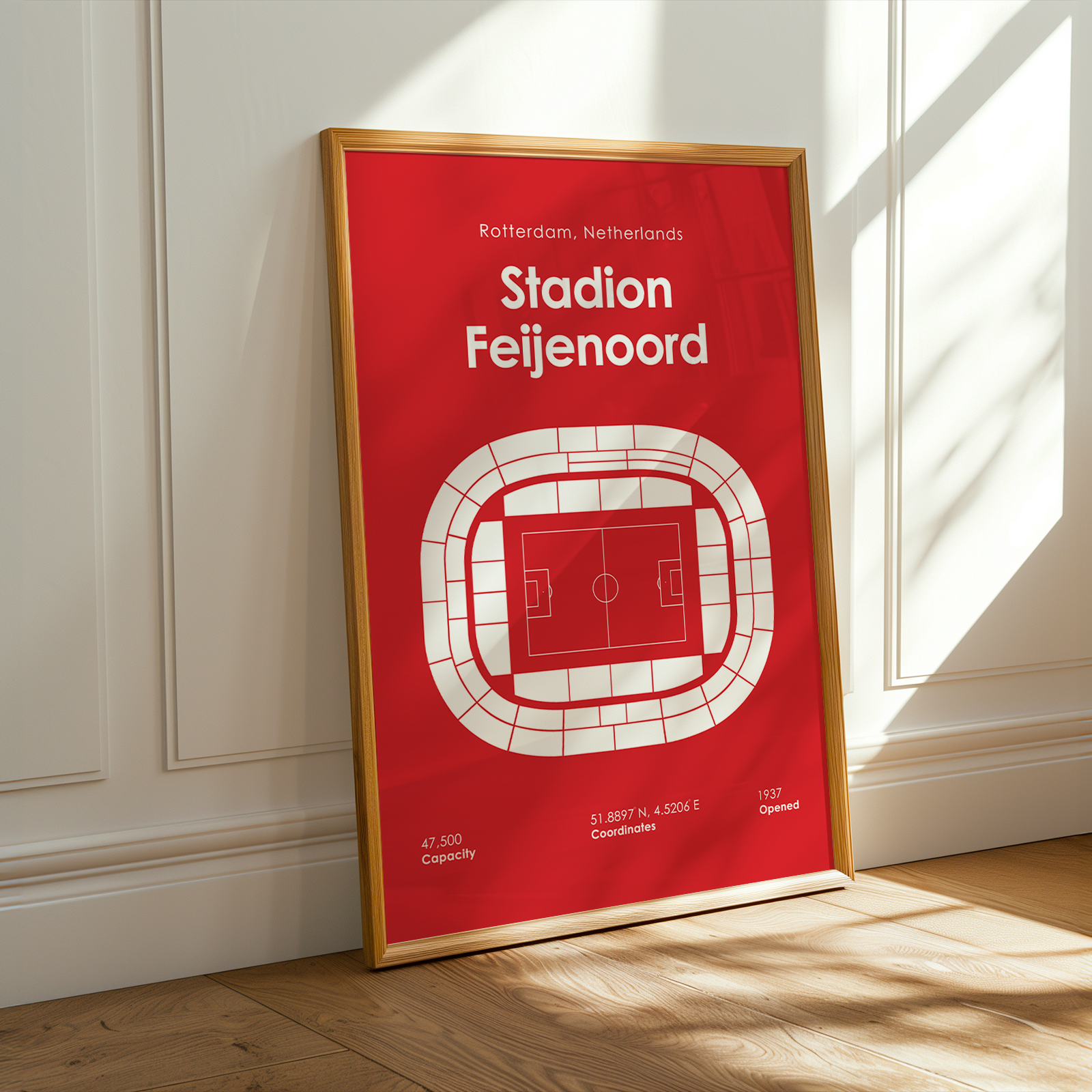 A framed red and white football stadium poster for Stadion Feijenoord, leaning against a white paneled wall on a light wood floor with warm sunlight streaming in from a window.