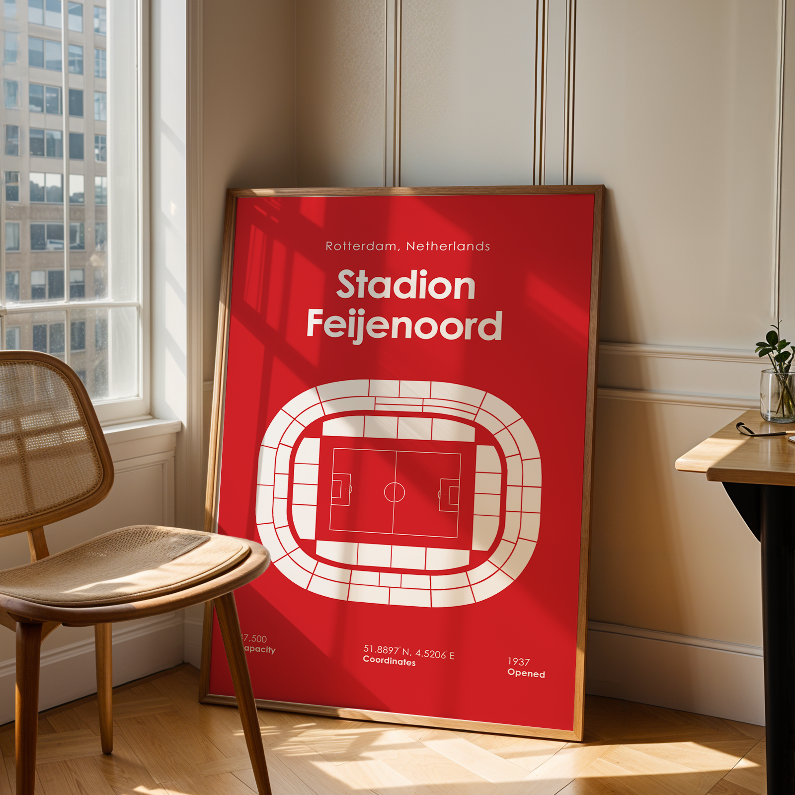 A framed red and white poster of Stadion Feijenoord stands on a wooden floor in a bright room next to a chair and a desk.