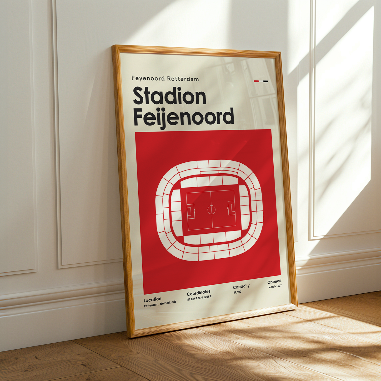 A framed poster of Feyenoord Rotterdam's Stadion Feijenoord, leaning against a white paneled wall on a wooden floor with warm sunlight streaming in.