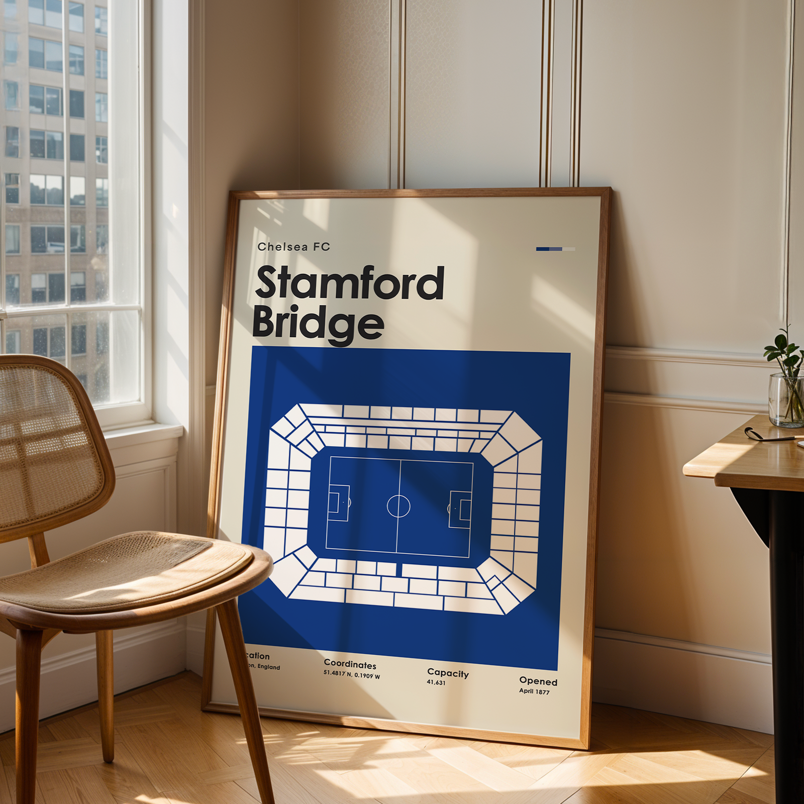 Framed poster of Stamford Bridge stadium in a room with a chair and window.