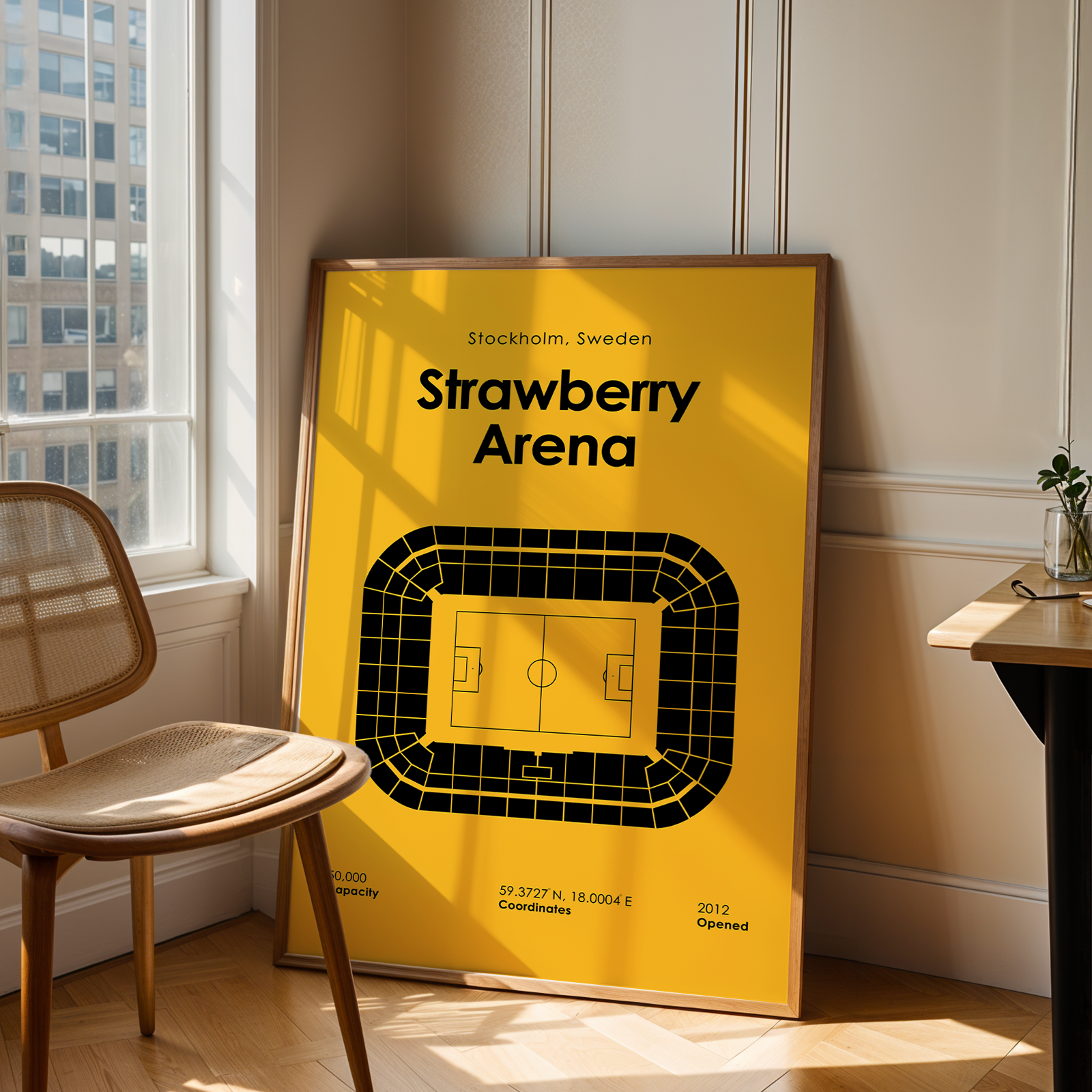 Framed poster of Strawberry Arena in Stockholm, Sweden, leaning against a wall in a room with a chair and table.