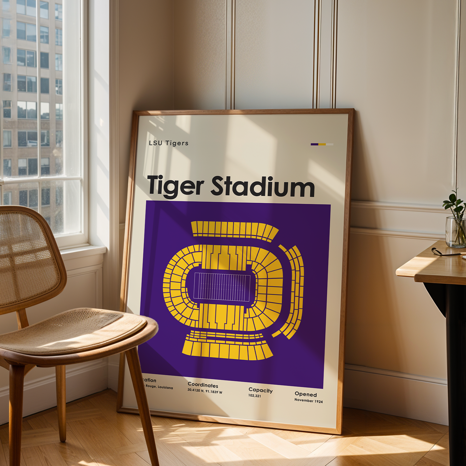 A framed LSU Tigers Tiger Stadium poster leaning against a wall in a sunlit room, next to a wooden chair and a small desk with a plant.