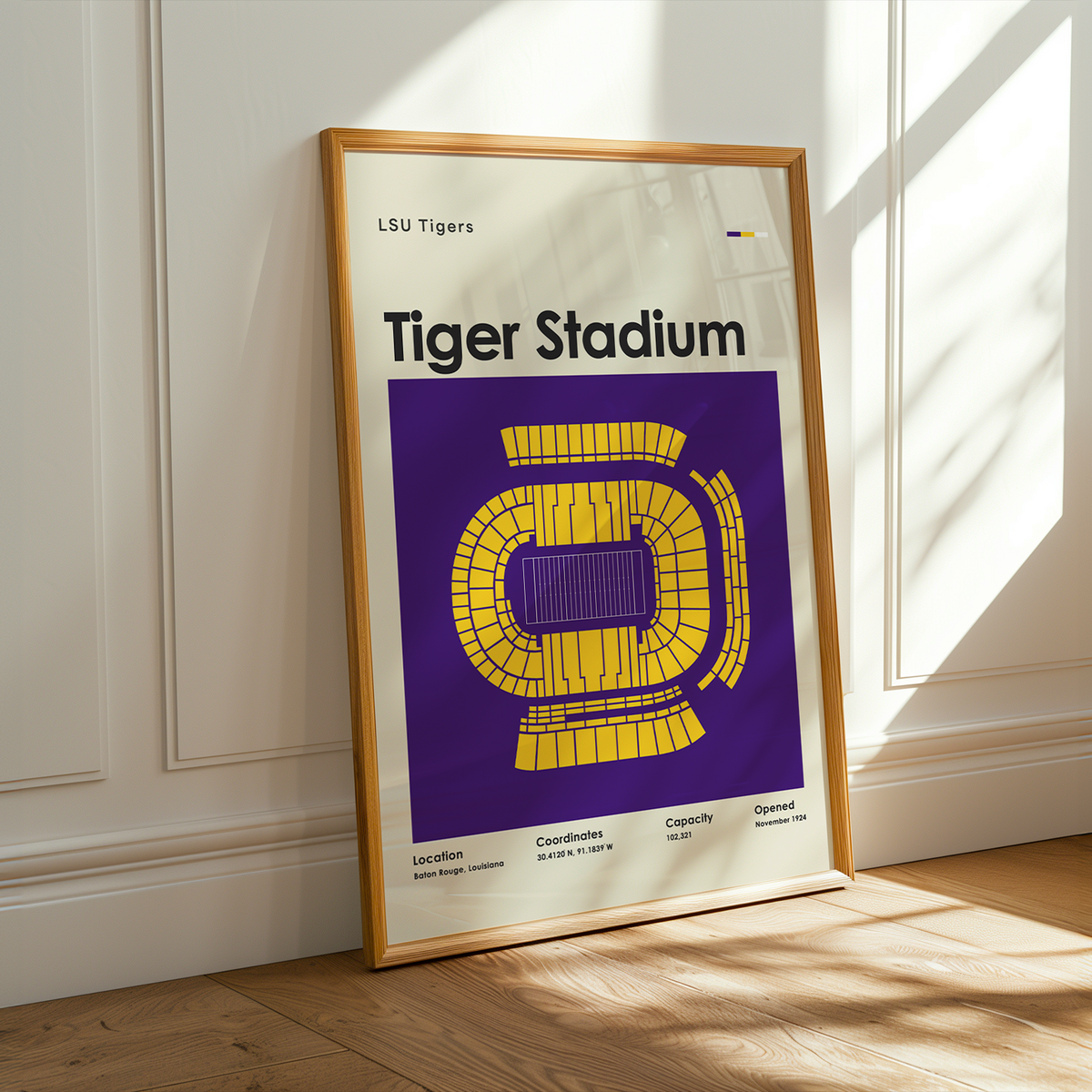 A framed LSU Tigers Tiger Stadium poster leaning against a white paneled wall on a light wood floor, with warm sunlight casting shadows across the wall and floor.