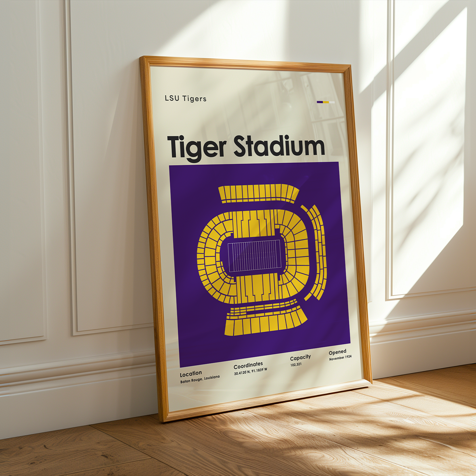 A framed LSU Tigers Tiger Stadium poster leaning against a white paneled wall on a light wood floor, with warm sunlight casting shadows across the wall and floor.