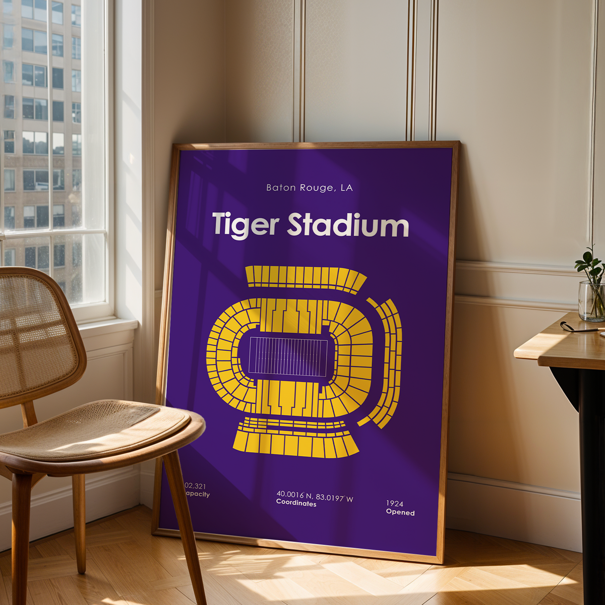 A framed poster of Tiger Stadium football game in Baton Rouge, LA, leaning against a wall in a modern room with a wooden chair and desk.