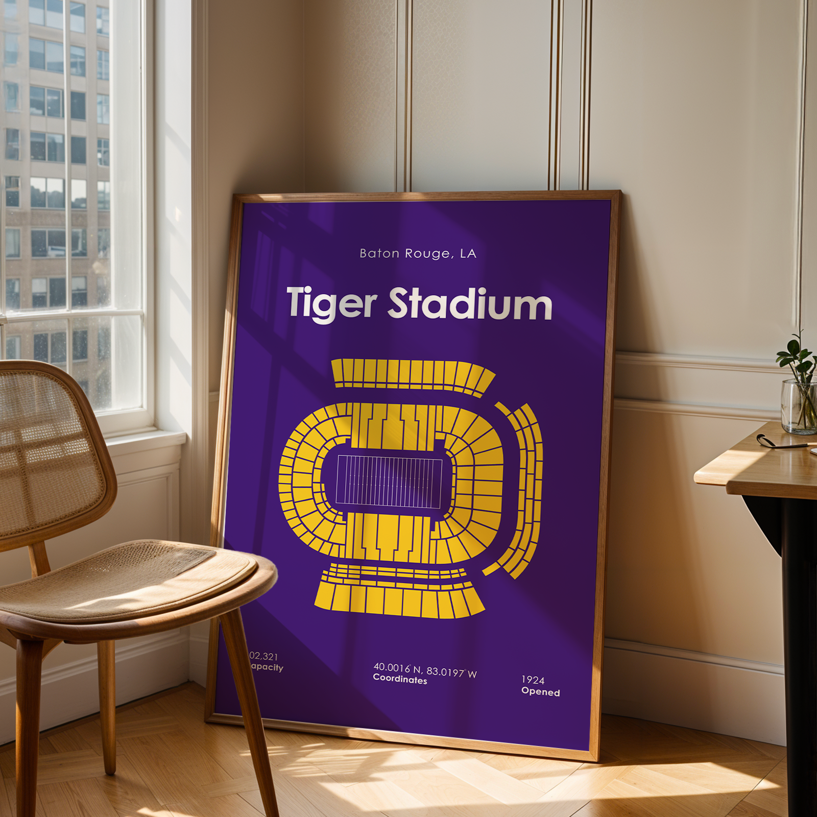 A framed poster of Tiger Stadium football game in Baton Rouge, LA, leaning against a wall in a modern room with a wooden chair and desk.