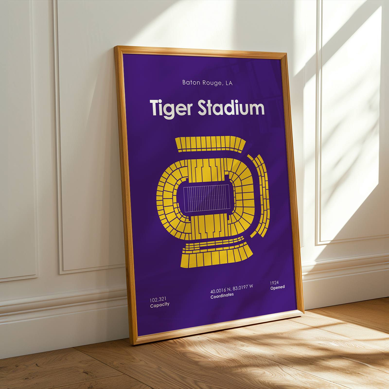A framed vertical poster of Tiger Stadium, leaning against a white paneled wall on a wooden floor. The poster features a purple background with a yellow stadium diagram and white text, enclosed in a light wood frame.