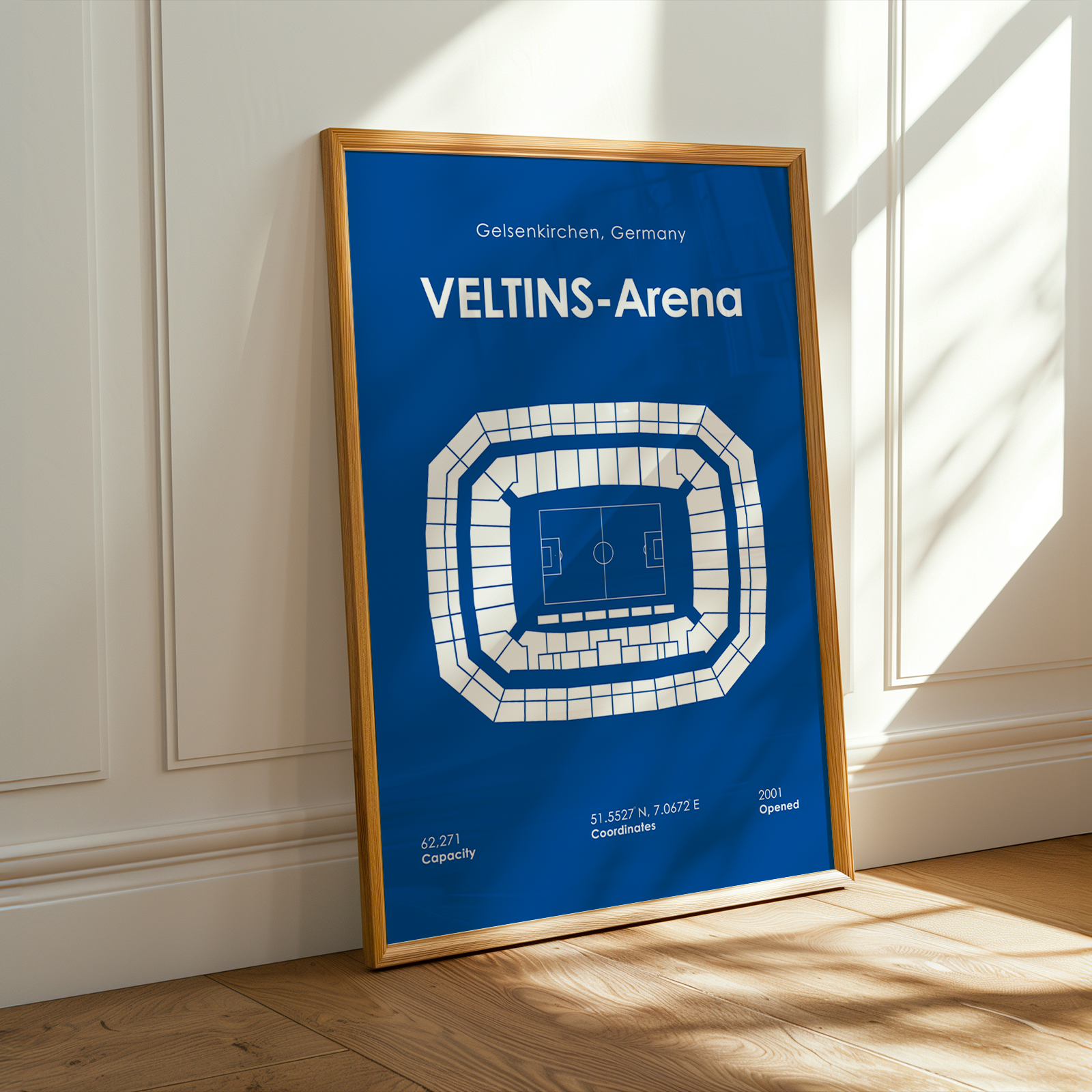 A framed blue sports poster of the Veltins-Arena in Gelsenkirchen, Germany, leaning against a white paneled wall on a wooden floor with warm sunlight and shadows.