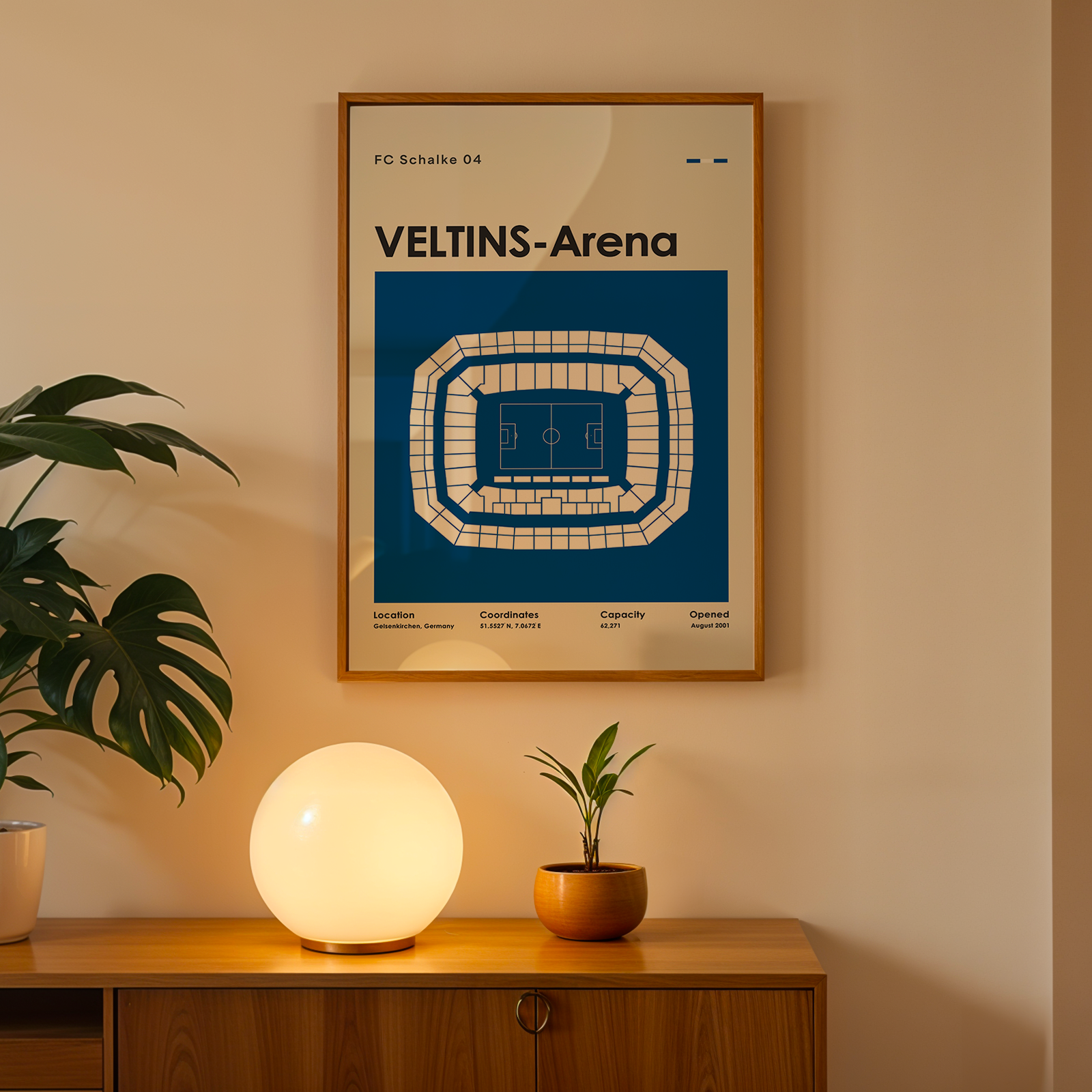 A framed poster of the Veltins-Arena football stadium in Leipzig, Germany, hanging on a beige wall above a wooden console table. The poster features a blue background with a white stadium layout and text.