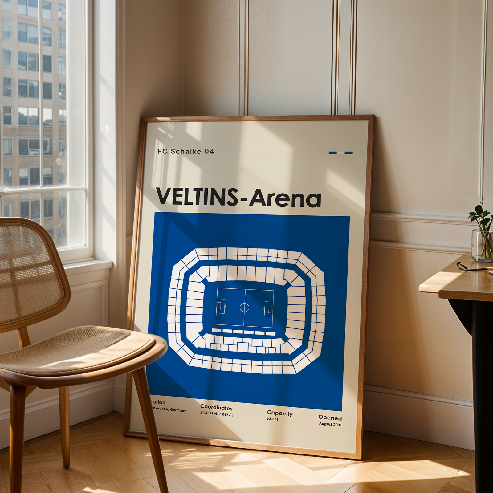 A framed poster of the Veltins-Arena football stadium in Leipzig, Germany, leaning against a cream paneled wall in a sunlit room next to a wooden chair and desk.