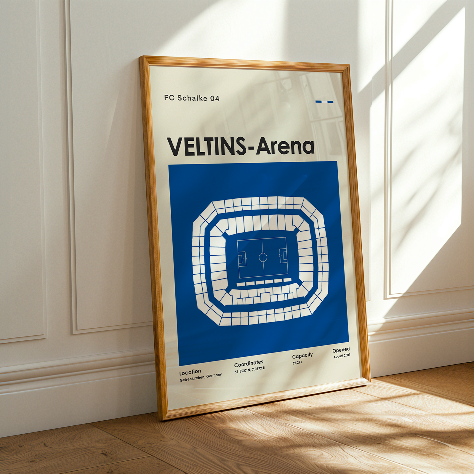 A framed sports poster of Veltins-Arena from FC Schalke 04 leaning against a white paneled wall on a wooden floor, with warm sunlight casting shadows.