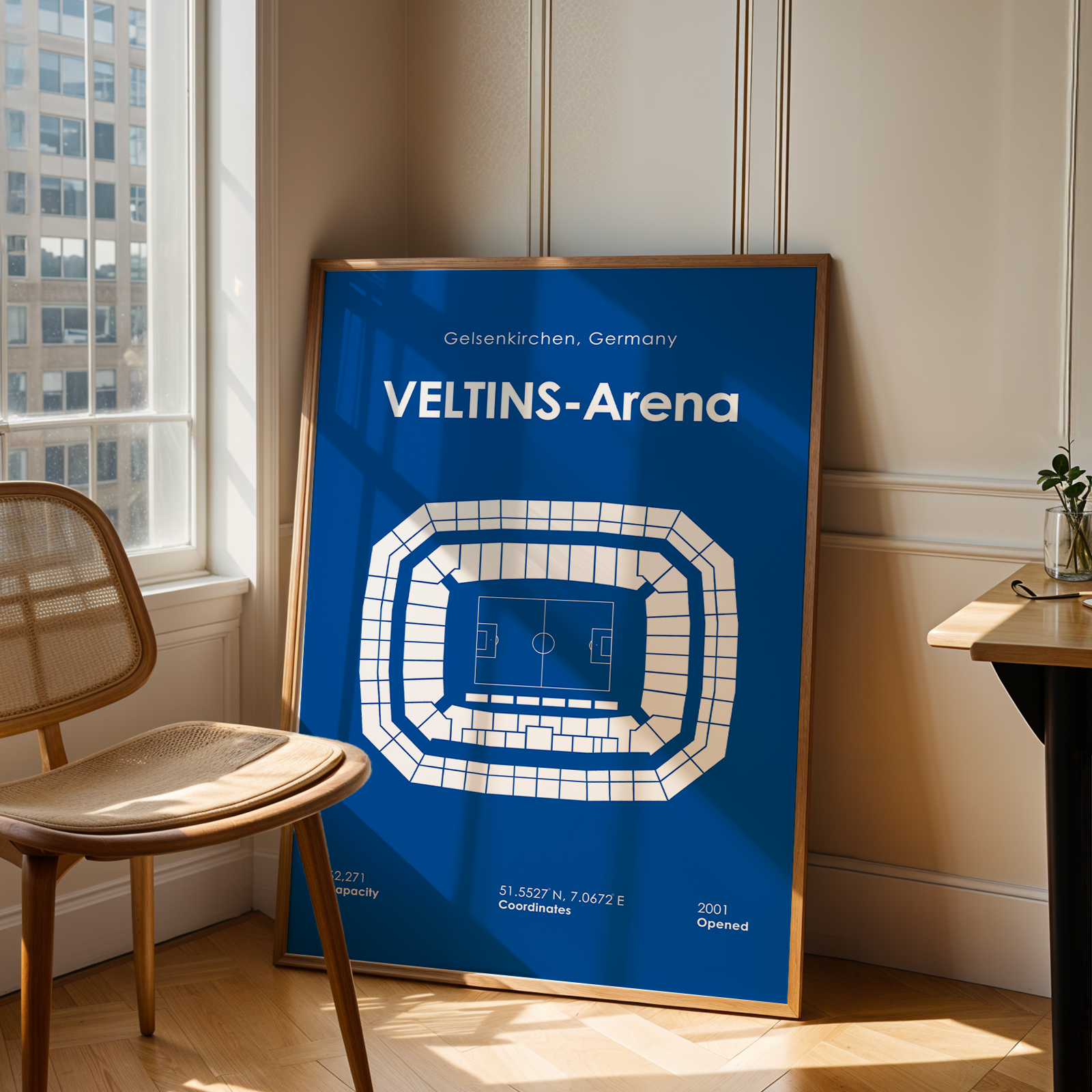 A framed blue and white Veltins-Arena sports poster leaning against a wall in a sunlit room with a wooden chair and desk visible.